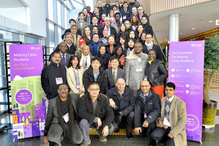 A large group of people poses for a photo on a staircase at an indoor conference, flanked by banners about making cities resilient.