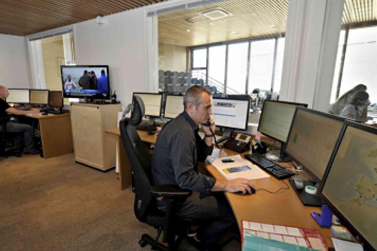 Several people work at desks with multiple computer monitors in a control room or office with large windows and wall-mounted screens.