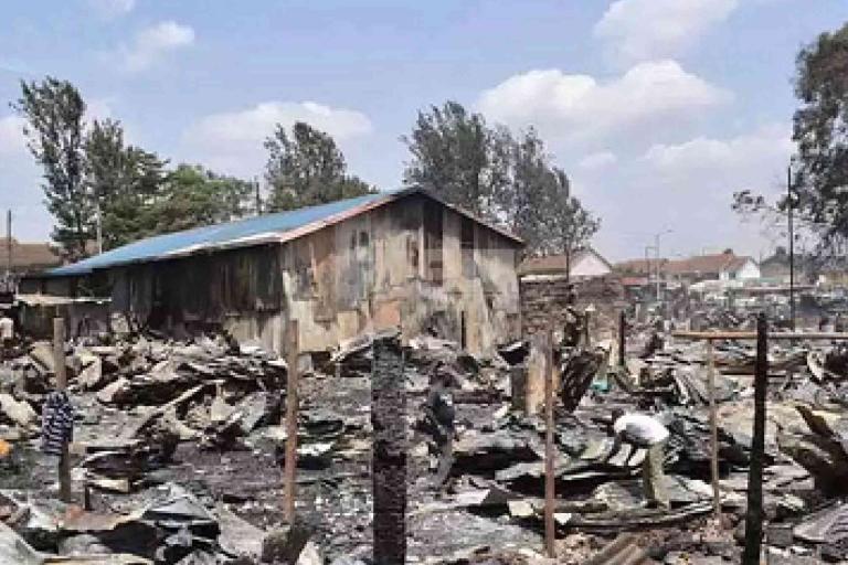 A corrugated metal building stands amid debris and charred remains of structures after a fire, with people inspecting the aftermath under a partly cloudy sky.