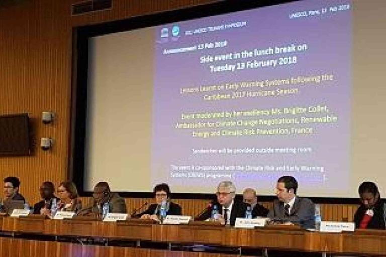 A panel of nine people sit at a long table with microphones during a side event at a conference, with a large presentation screen behind them displaying event details.