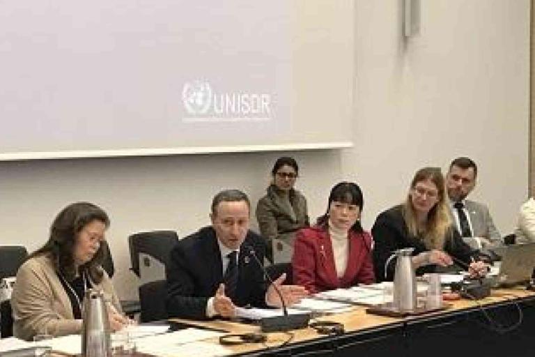 A panel of six people sits at a conference table with documents and microphones, with a UNISDR logo projected on the wall behind them.