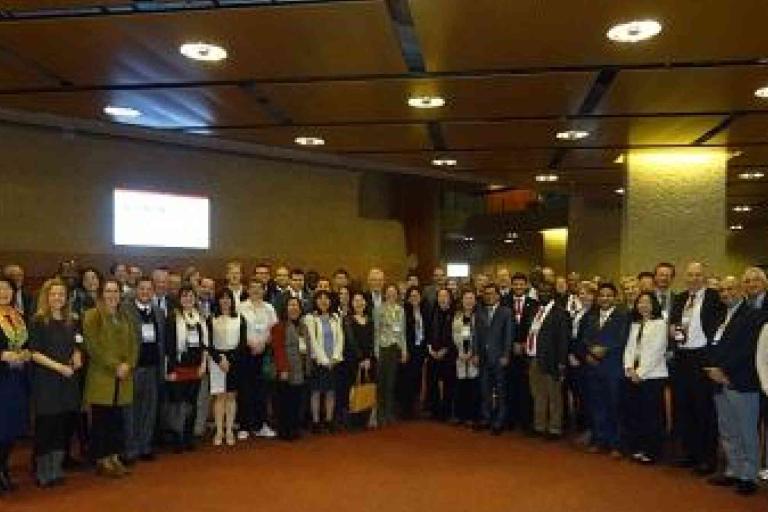 A large group of people stands indoors in a conference or event hall, posing for a group photo under a high ceiling with lights.