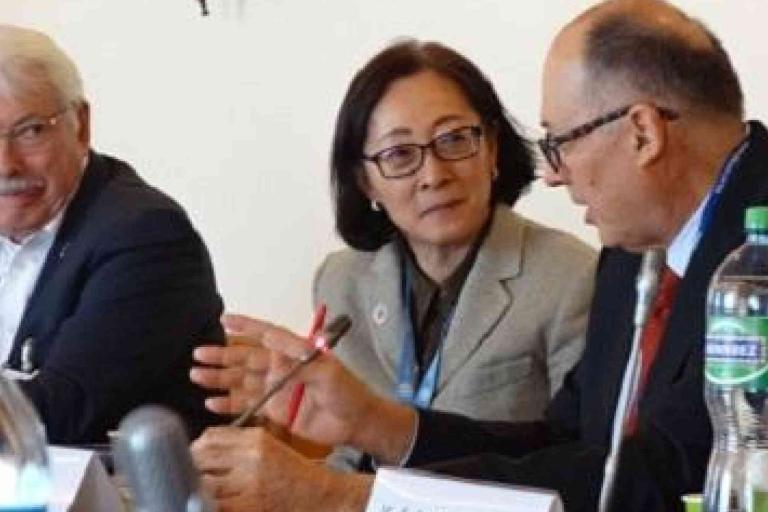 Three people in business attire sit at a conference table, engaged in conversation, with microphones and water bottles in view.
