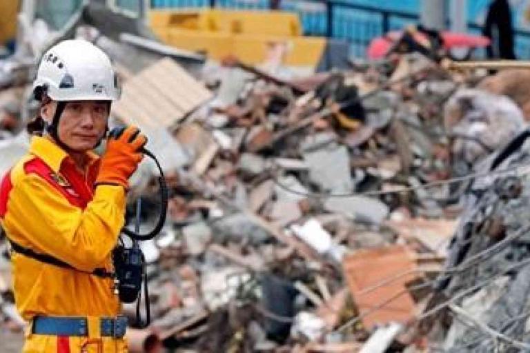 A rescue worker in a yellow uniform and helmet communicates on a radio while standing in front of a large pile of rubble.