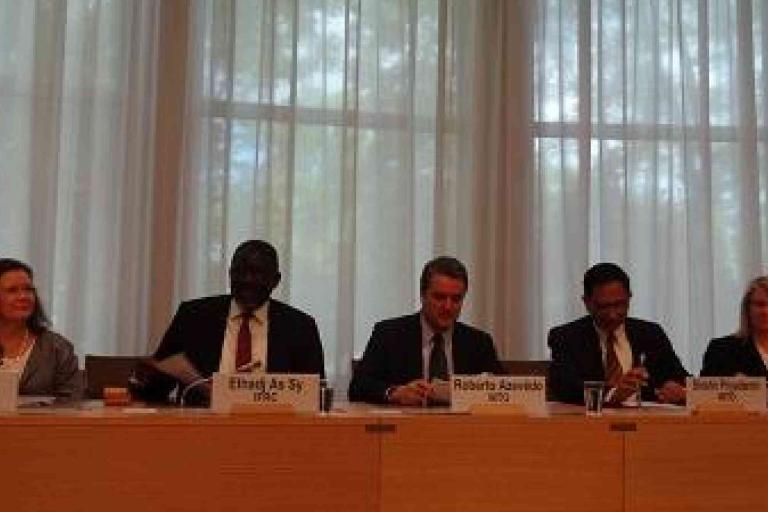 Five people sit behind a desk in a conference room, with nameplates in front of them and large windows with curtains in the background.