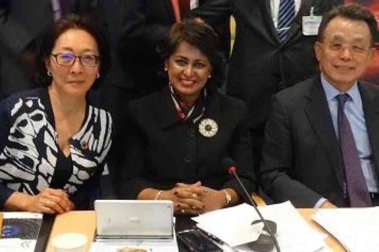Three people in business attire seated at a conference table with documents, a laptop, and microphones in front of them, posing for a group photo.