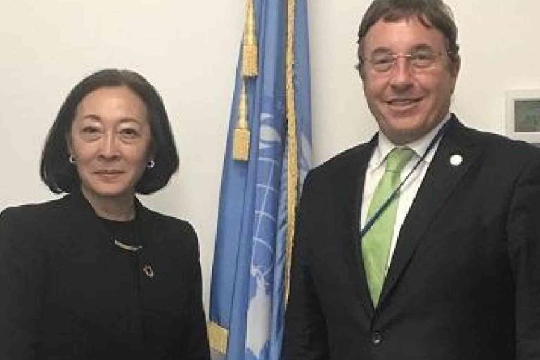 Two people in formal attire stand in front of a United Nations flag, posing for a photo indoors.