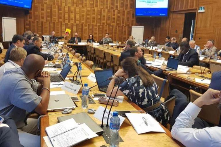 A group of people in business attire sit around a large conference table with laptops, papers, and microphones in a wood-paneled meeting room.