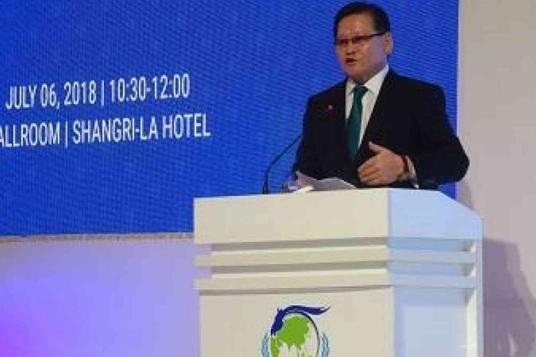 A man in a suit speaks at a podium during an event at Shangri-La Hotel, with a screen showing the date July 6, 2018, and event time 10:30-12:00.