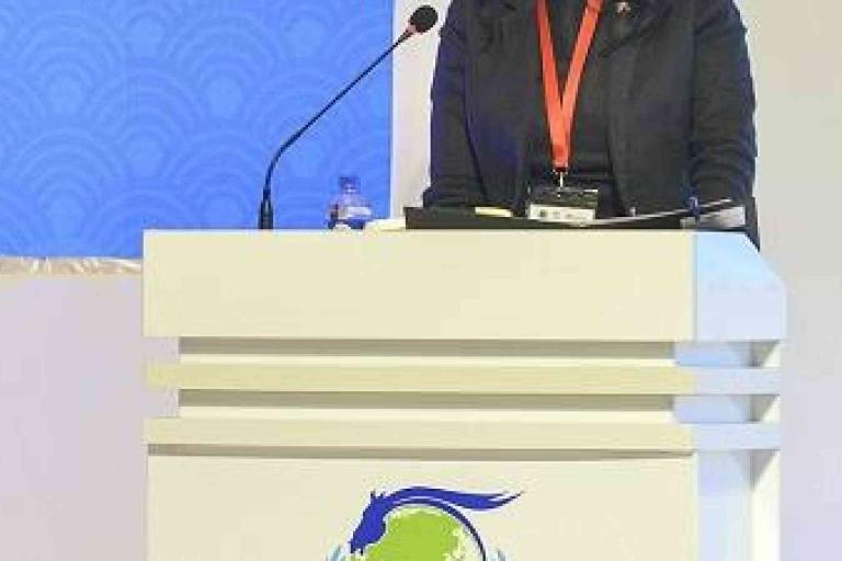 A person stands at a podium with a microphone, wearing a dark jacket and red lanyard. The podium features a blue and green logo with a horse.