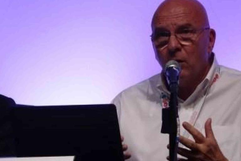 Bald man in glasses speaks into a microphone at a panel discussion, seated at a table with a laptop and water bottles.
