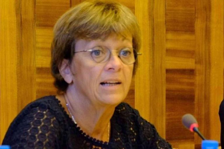 A woman with short brown hair and glasses speaks into a microphone at a wooden-panel table.