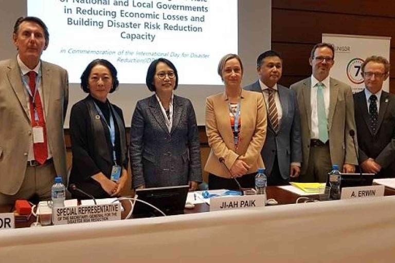 A group of seven professionals stand in front of a presentation screen at a conference on disaster risk reduction.