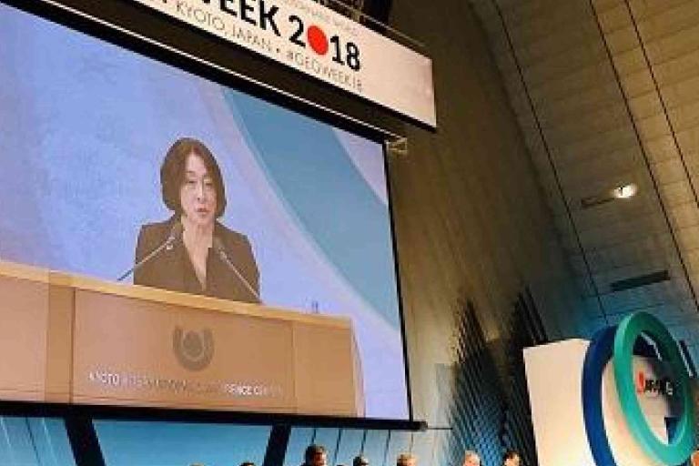 A woman speaks at a podium during a conference, with "WEEK 2018" and "Kyoto, Japan" displayed on a large screen above her. Several people are seated on stage.