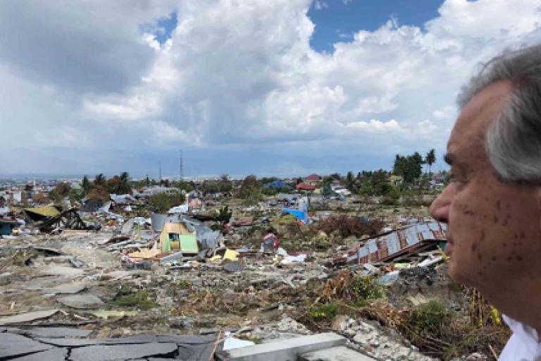 A man looks over a landscape of destroyed buildings and debris under a cloudy sky, indicating the aftermath of a natural disaster.