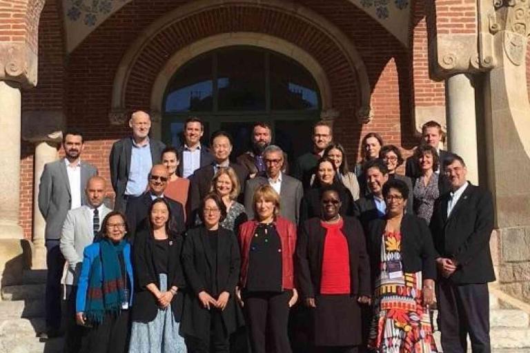 A group of people pose for a photo on the steps of a historic brick building with arched windows and ornate pillars.