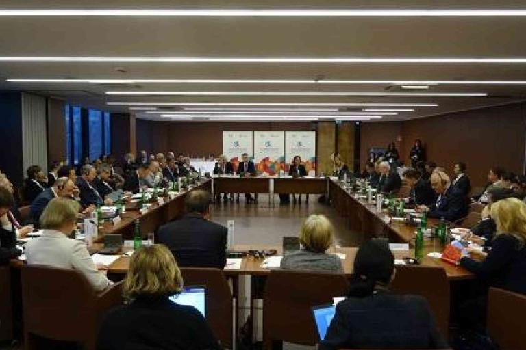 People sit around a large rectangular conference table in a modern meeting room, engaged in a formal discussion.