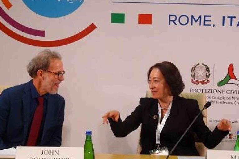Two people sit at a conference table in front of a backdrop with logos and "Rome, Italy" text, engaged in conversation. Bottled water and nameplates are visible.