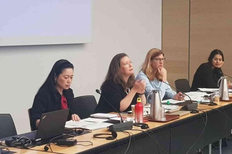 Four women sit at a conference table with laptops, documents, and microphones, appearing engaged in a meeting or discussion.