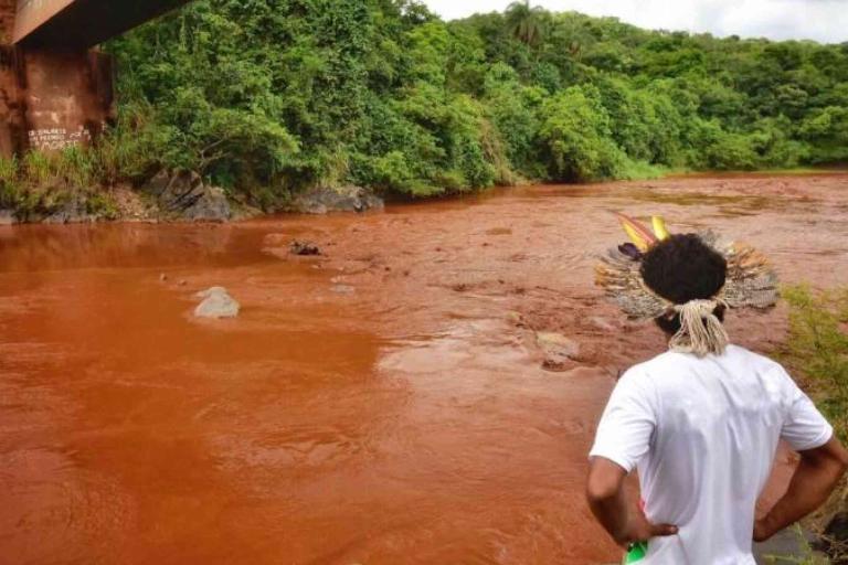 A person wearing a feathered headdress looks at a reddish-brown river beneath a bridge, with dense green vegetation on both banks.