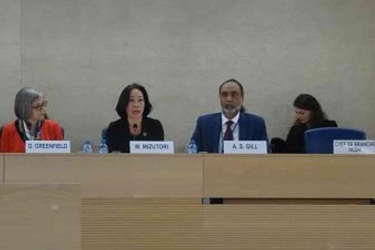 Four panelists sit at a conference table with microphones, nameplates, and water bottles; one person is speaking while others listen.
