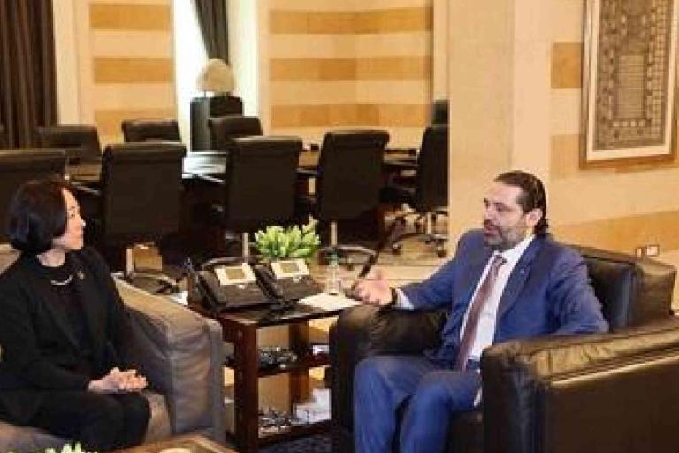 Two people sit across from each other in an office lounge area, engaged in conversation. Office chairs and tables are visible in the background.