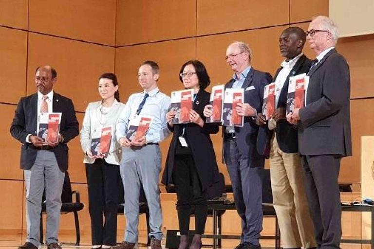Seven people stand in a row on stage holding documents, in front of a podium with a United Nations emblem in a wood-paneled room.
