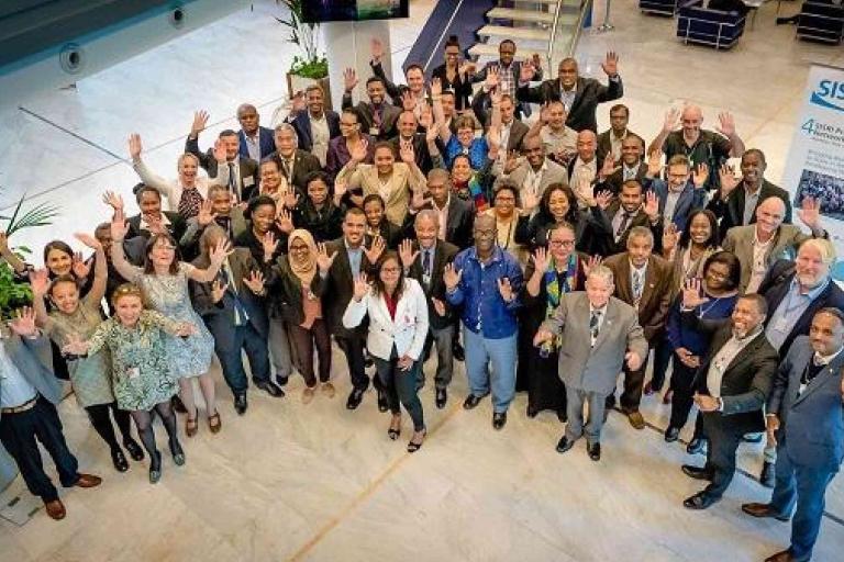 A large group of professionally dressed people stand indoors, smiling and waving at the camera. A SISR banner is visible in the background.