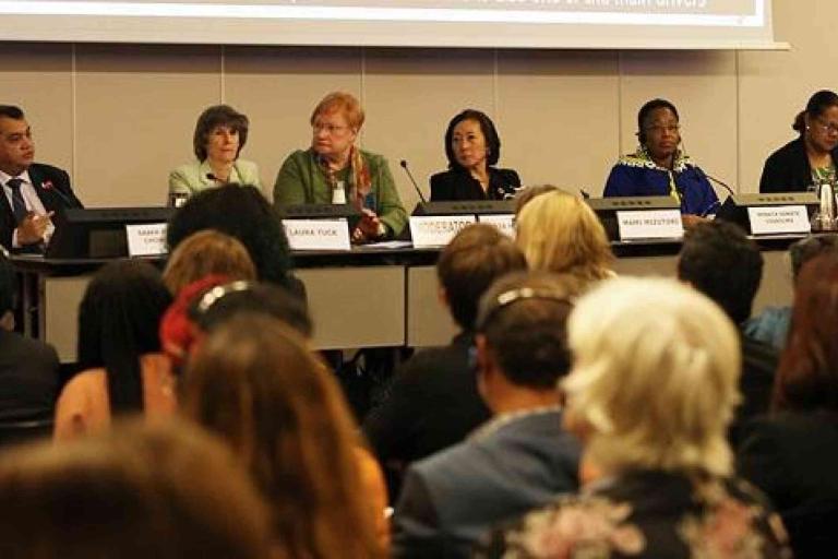 A panel of six people sit at a long table with microphones, addressing an audience at a conference or formal meeting.
