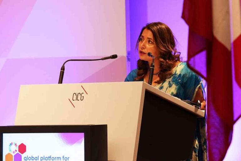 A woman stands at a podium labeled "CICG," speaking into a microphone at a conference with a display screen and flags in the background.