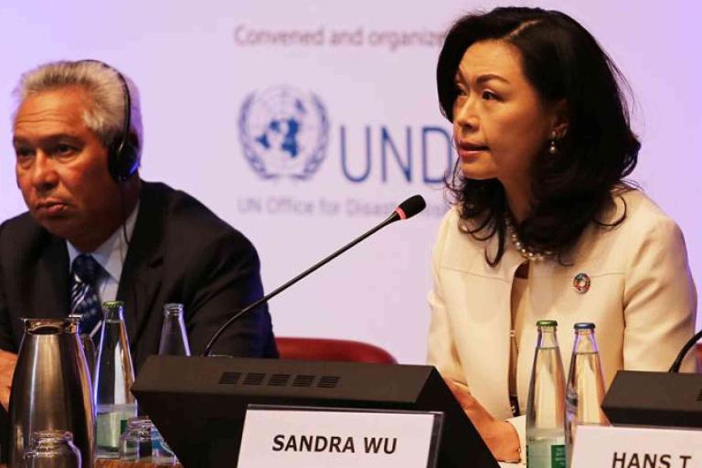 Two individuals sit at a conference table with microphones and nameplates; Sandra Wu is speaking, with a UN Office for Disaster Risk Reduction logo in the background.