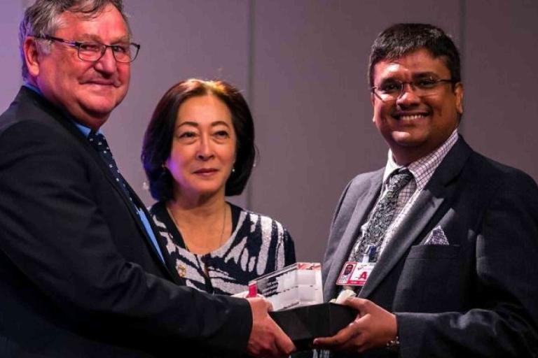 Three people in formal attire stand together; two men shake hands while holding a boxed item, with a woman standing between them, indoors against a plain background.