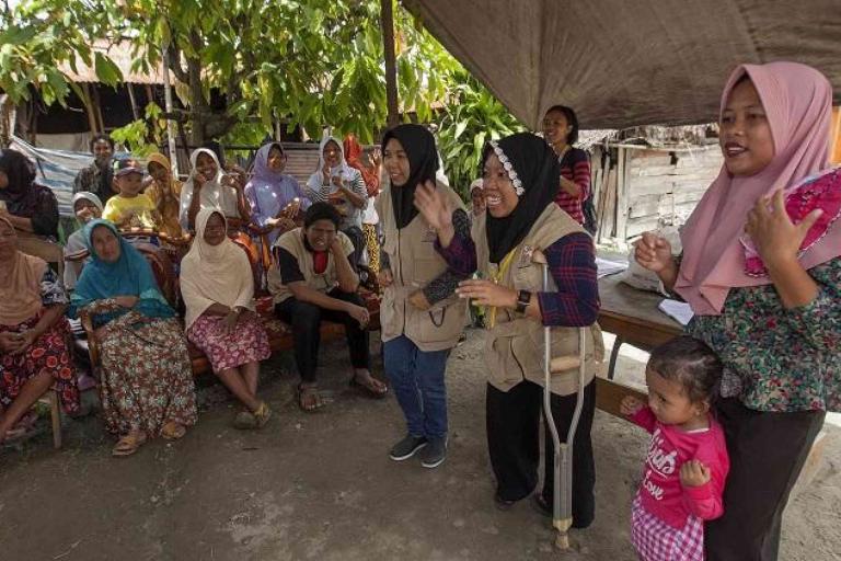 A group of women and children gather outdoors under a canopy, with some standing and speaking while others sit and listen.