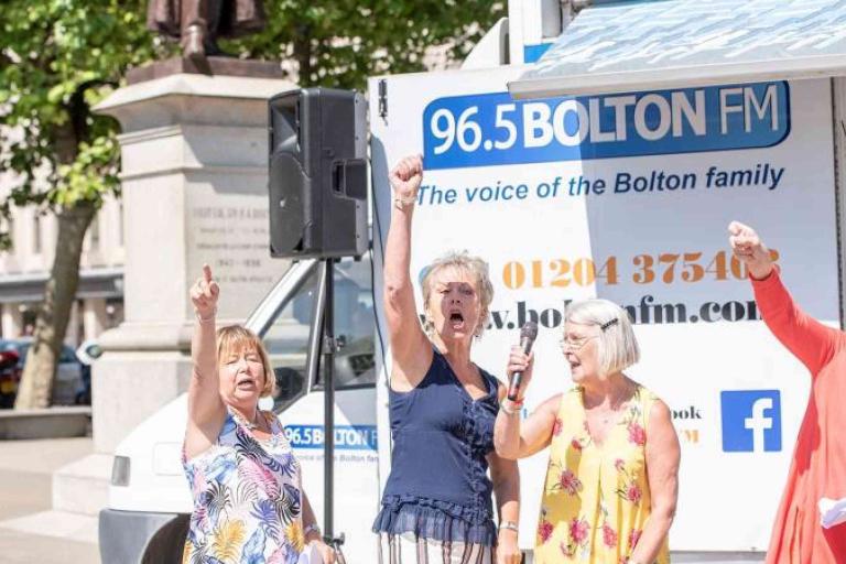 Four women stand and gesture energetically in front of a 96.5 Bolton FM radio van during an outdoor event.
