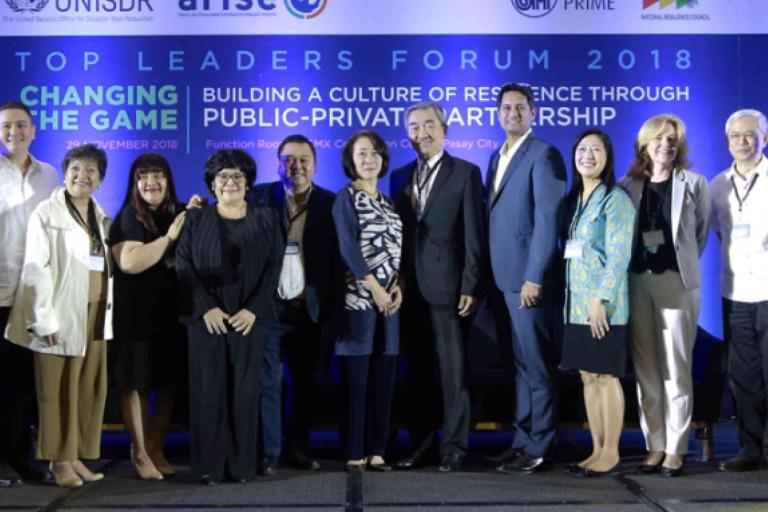 A group of people in business attire stands on stage in front of a blue banner that reads "Top Leaders Forum 2018: Building a Culture of Resilience Through Public-Private Partnership.