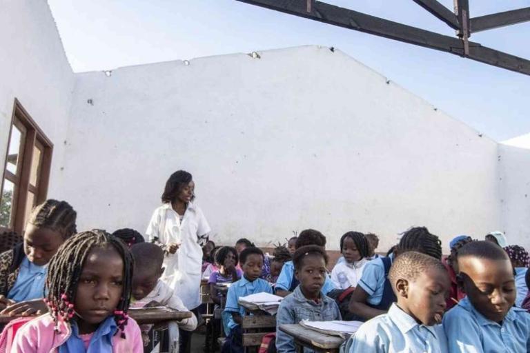 A teacher stands in front of students seated at desks in a partially roofless classroom with white walls, under daylight.