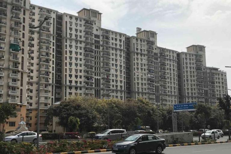 A multi-storey residential apartment building with balconies stands beside a busy road lined with trees and parked cars on a cloudy day.