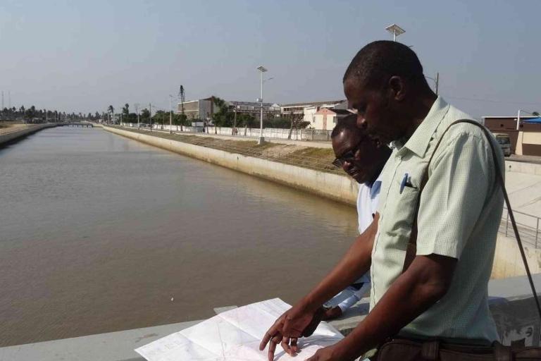 Two men stand on a bridge over a canal, examining documents or plans, with buildings and streetlights visible in the background.
