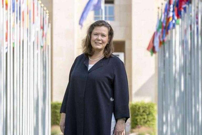 A woman stands outdoors between rows of flagpoles displaying various international flags, with a building and greenery in the background.