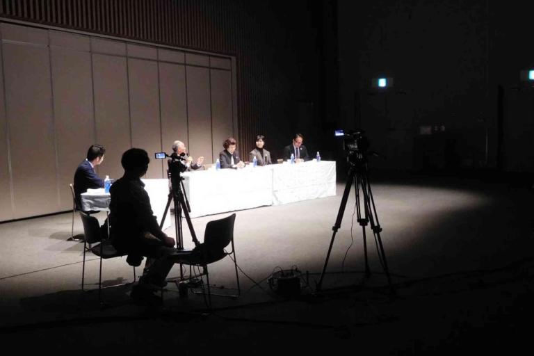 A panel of four people sits at a table with microphones under spotlights, while a camera operator films and another person sits nearby in a dimly lit room.