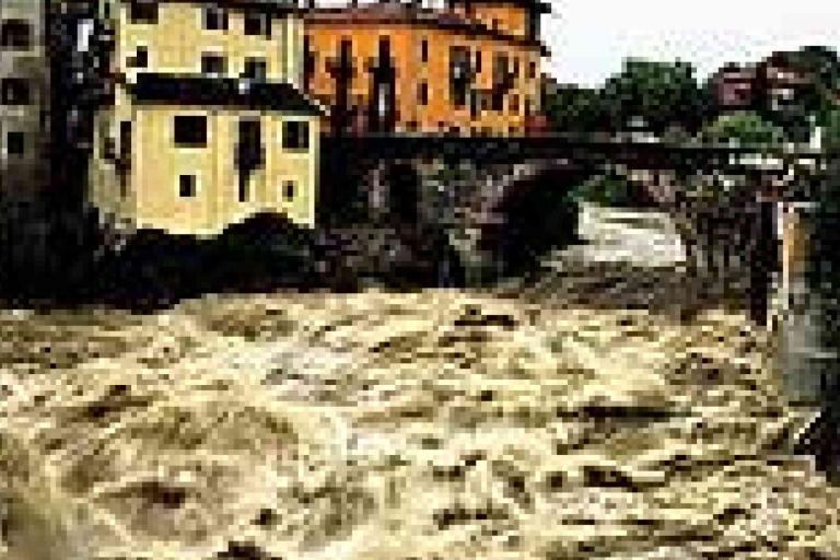 Floodwaters surge through a river in a town, with turbulent water nearly reaching buildings and a bridge in the background.