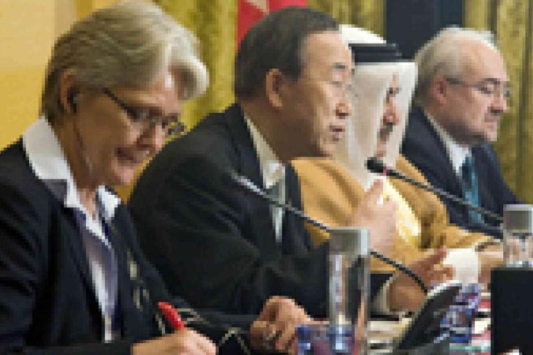 Four people in formal attire sit at a conference table with microphones and papers, participating in a panel discussion or diplomatic meeting.
