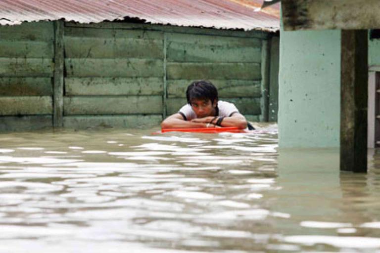 A person clings to a floating object in deep floodwater beside a wooden fence and a building.