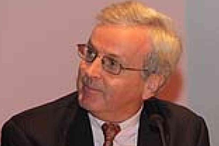 An older man wearing glasses, a suit, and a tie is looking to his left against a plain background.