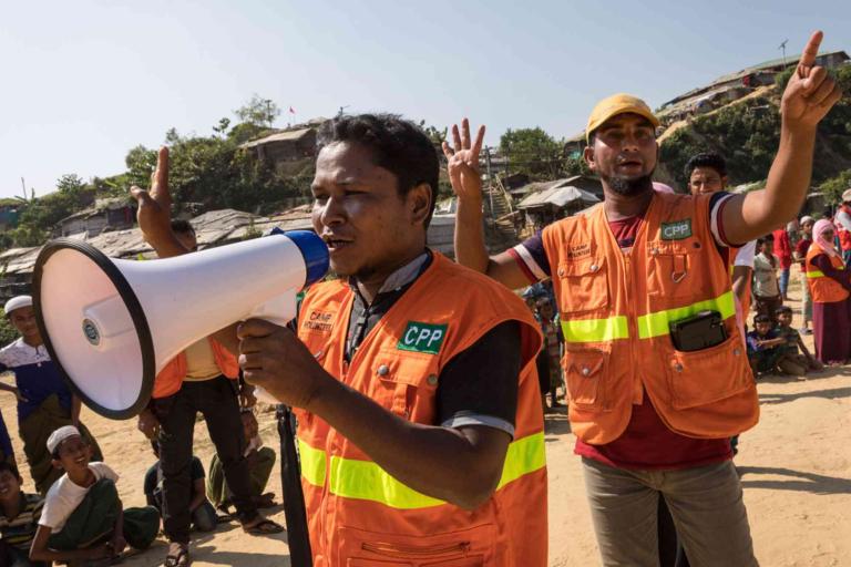 Two men in orange vests use a megaphone and hand signals to address a crowd outdoors, with several people seated and some buildings in the background.