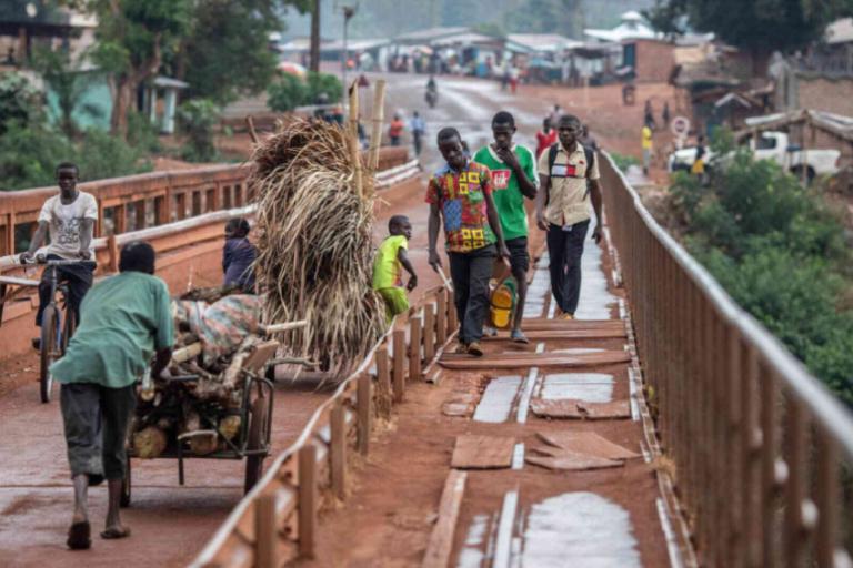 People walk and push carts across a narrow, rustic bridge in a rural area, with buildings and greenery visible in the background.