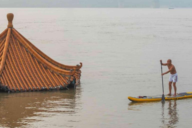 A man paddleboards past the partially submerged roof of a traditional building in floodwaters.