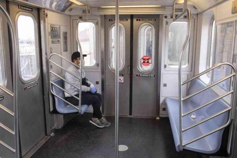 A person wearing a mask and gloves sits alone on a mostly empty subway train.