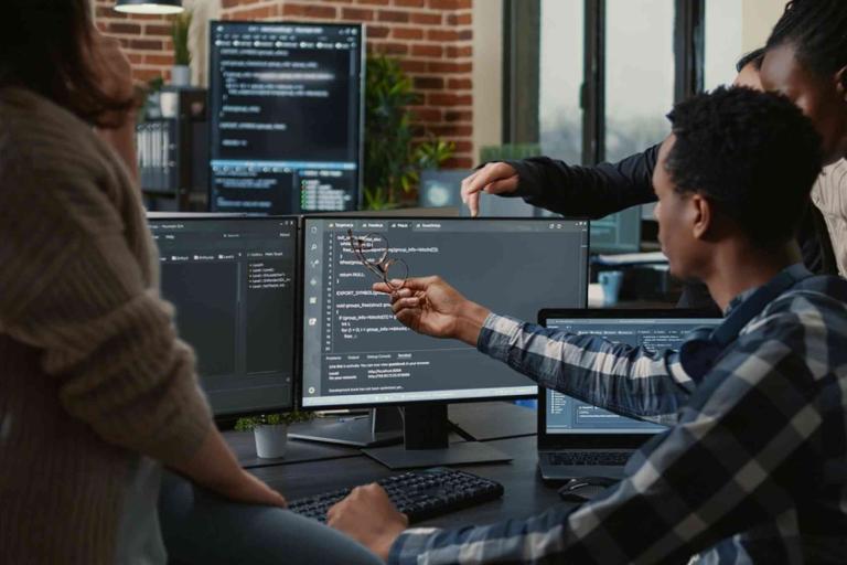 Group of people collaborating at a desk with multiple computer monitors displaying code in a modern office setting.