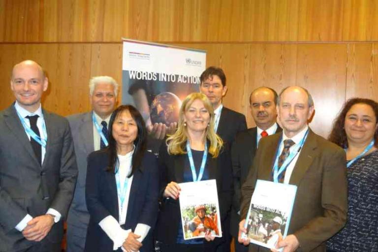 Eight people in business attire stand together indoors, some holding documents, in front of a wooden wall and a banner that reads "WORDS INTO ACTION.